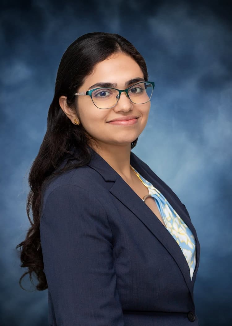 A professional woman with long, dark hair and glasses, dressed in a navy blazer over a patterned blouse. She stands against a blue background, smiling confidently, embodying the expertise and compassion essential in the field of nephrology.