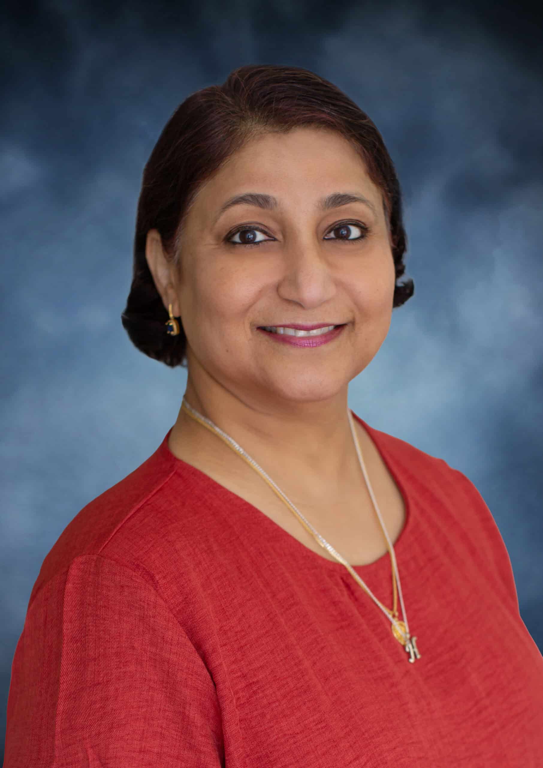 Parveen Naaz, M.D., with short dark hair, wearing a red top and gold jewelry, smiles at the camera against a blue gradient background.
