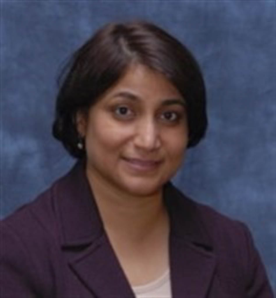 Parveen Naaz-Ikramuddin, M.D., with short dark hair, wearing a dark blazer over a light top, sits in front of a blue background and smiles slightly at the camera.