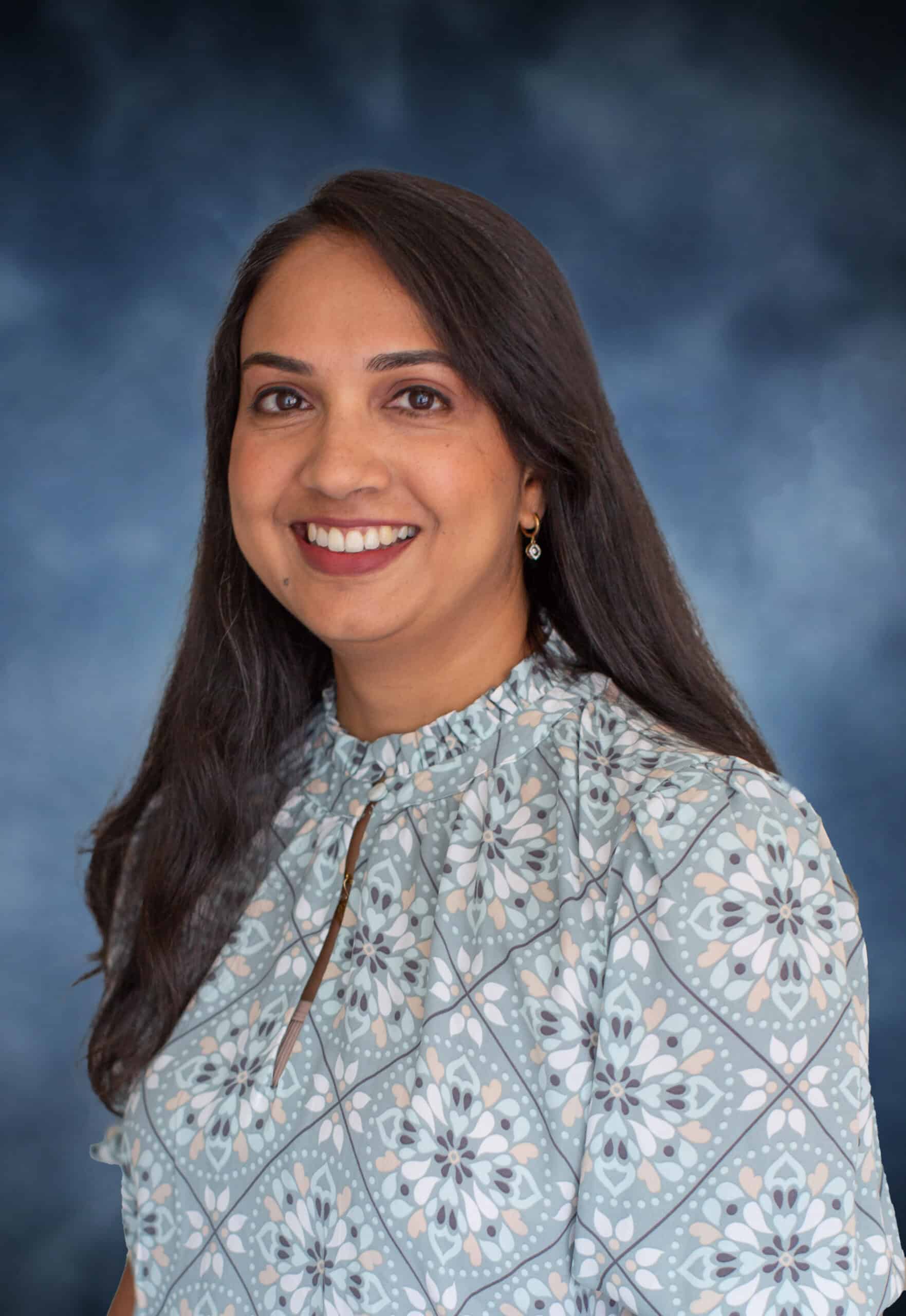 A woman with long dark hair and a patterned blouse smiles in front of a blue, softly blurred background.