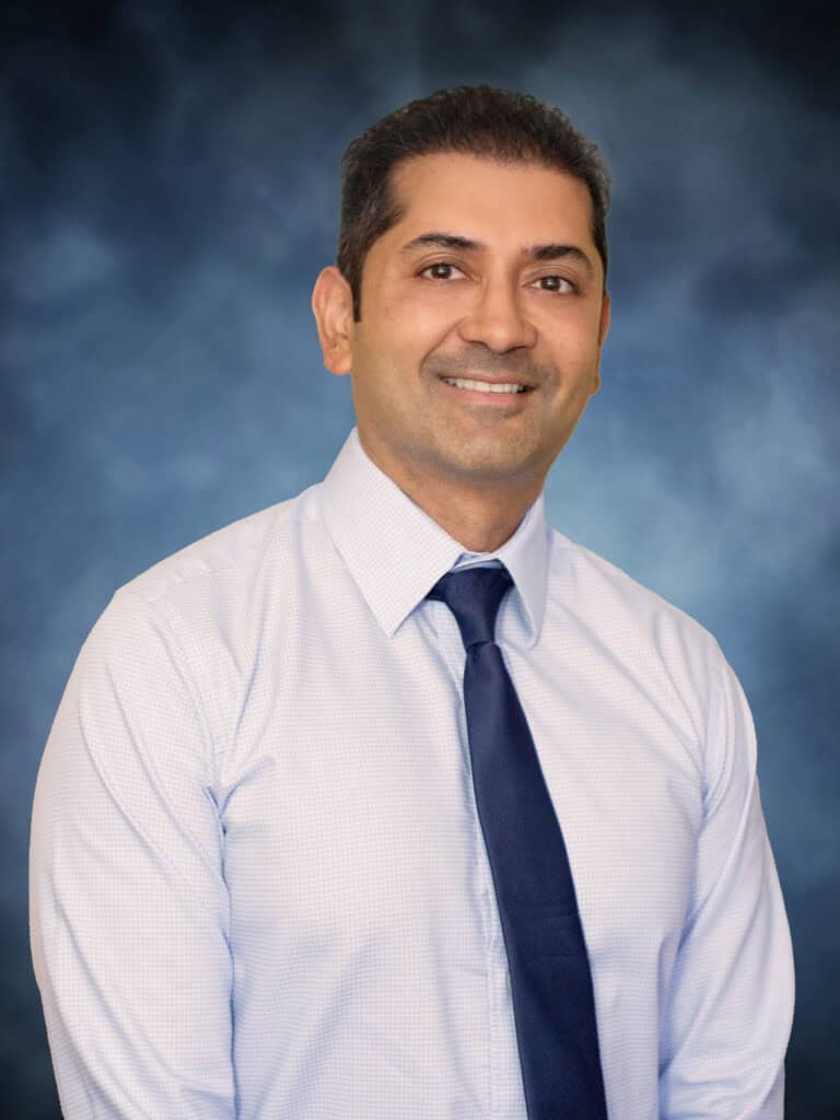 A man wearing a light-colored dress shirt and dark tie smiles at the camera against a blue, softly blurred background. This is Dr. Chaitanya Desai, M.D.
