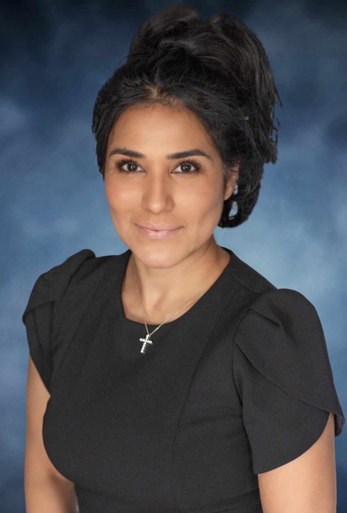 Dr. Anjali Mathew, with dark hair in an updo, wearing a black dress with puffed sleeves and a silver cross necklace, poses against a blue and gray gradient background, smiling softly at the camera for RenalCare.