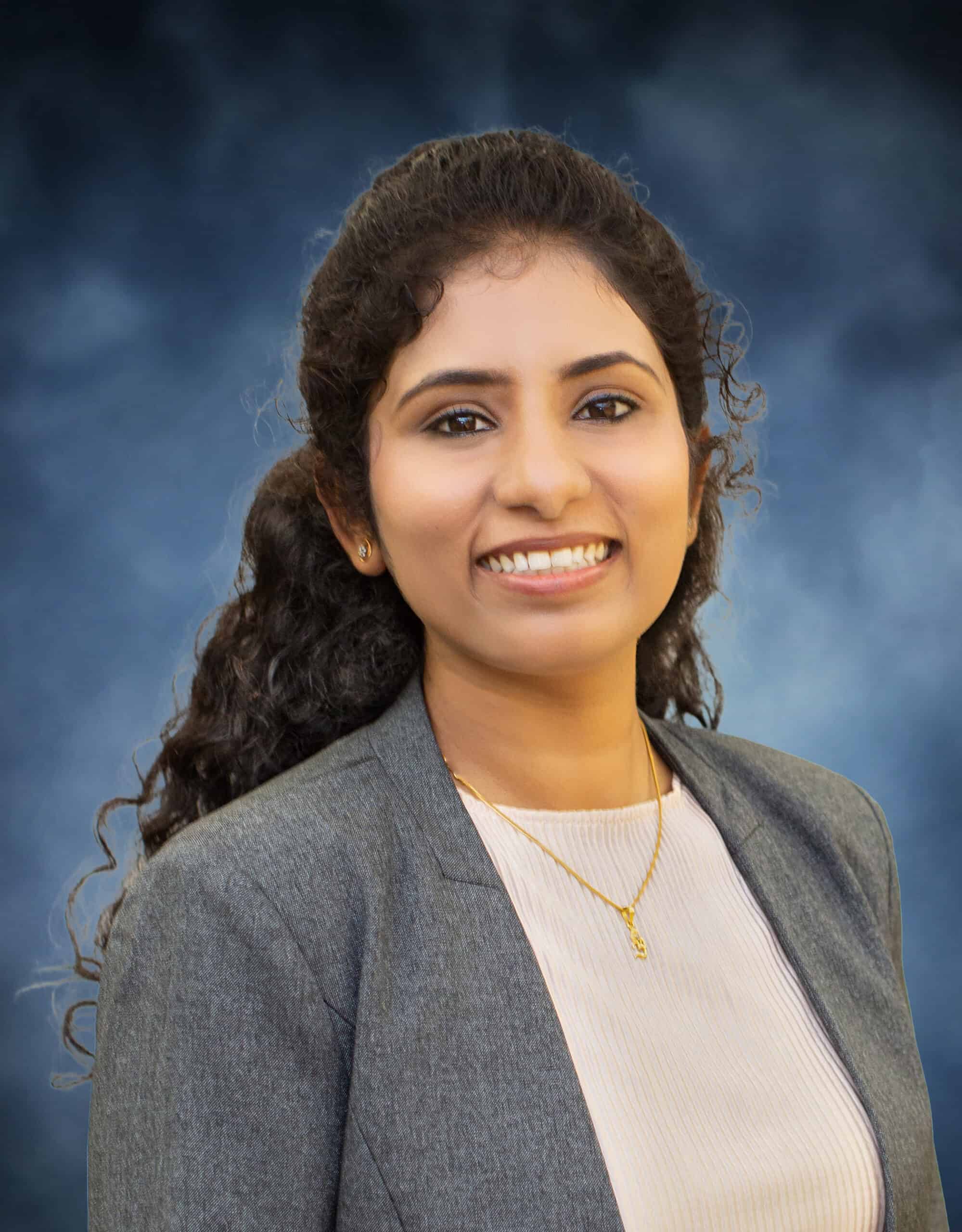A woman with long curly hair, wearing a gray blazer over a light top and a gold necklace, smiles in front of a blue, softly blurred background—Krishna Van Nemalidinne.