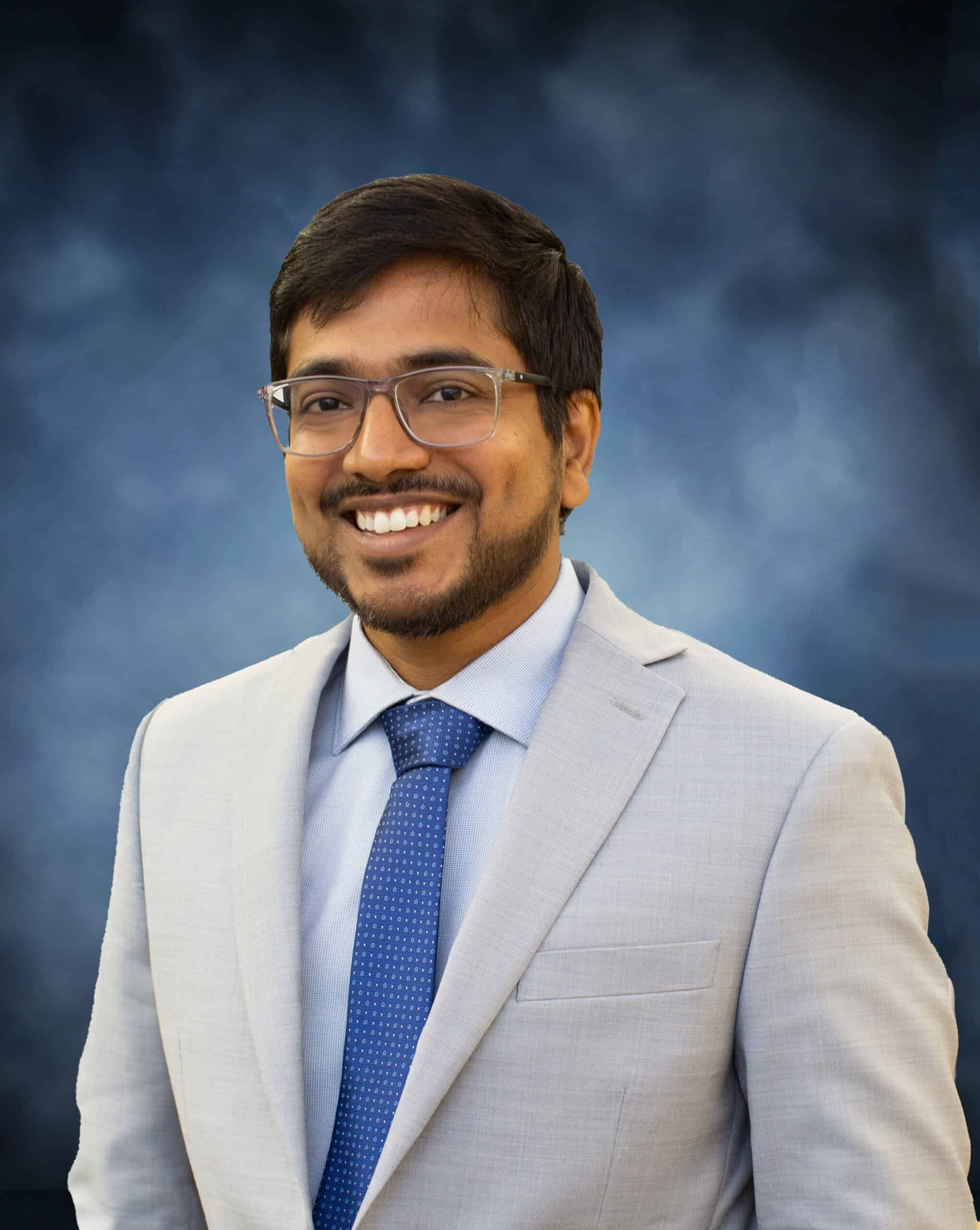 Abinay Vanteru, a man wearing glasses, a light gray suit, blue shirt, and blue polka dot tie, stands smiling in front of a blurred blue background.