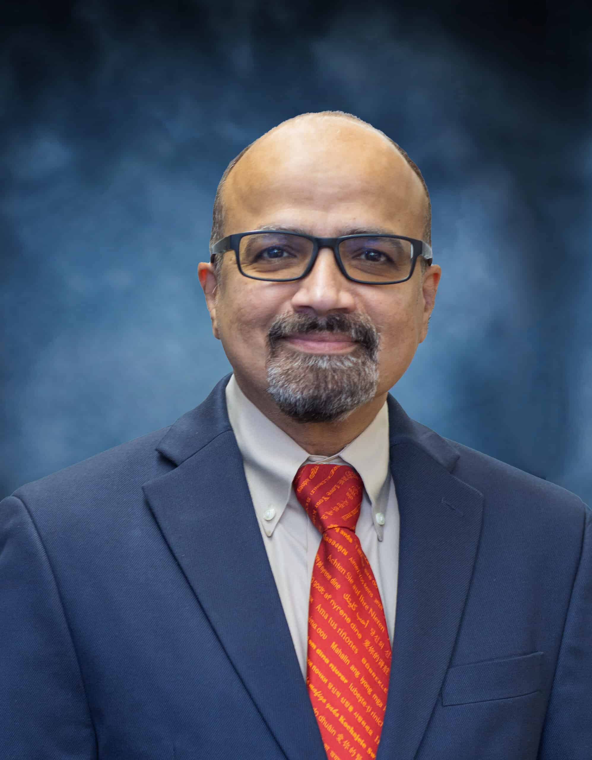 A bald man with glasses, a goatee, and mustache—Vish Halukurike, M.D.—wearing a dark blue suit, light shirt, and red tie, stands in front of a blue textured backdrop, smiling slightly at the camera.