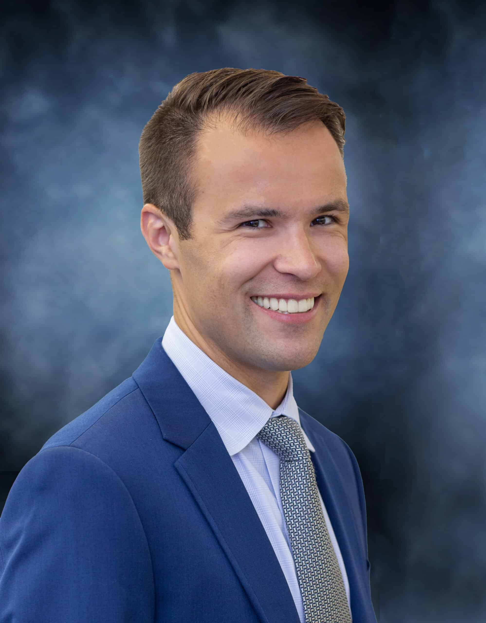 A man in a blue suit and tie smiles at the camera against a dark, blurred background, radiating the professional confidence of Ian Lewis, M.D.