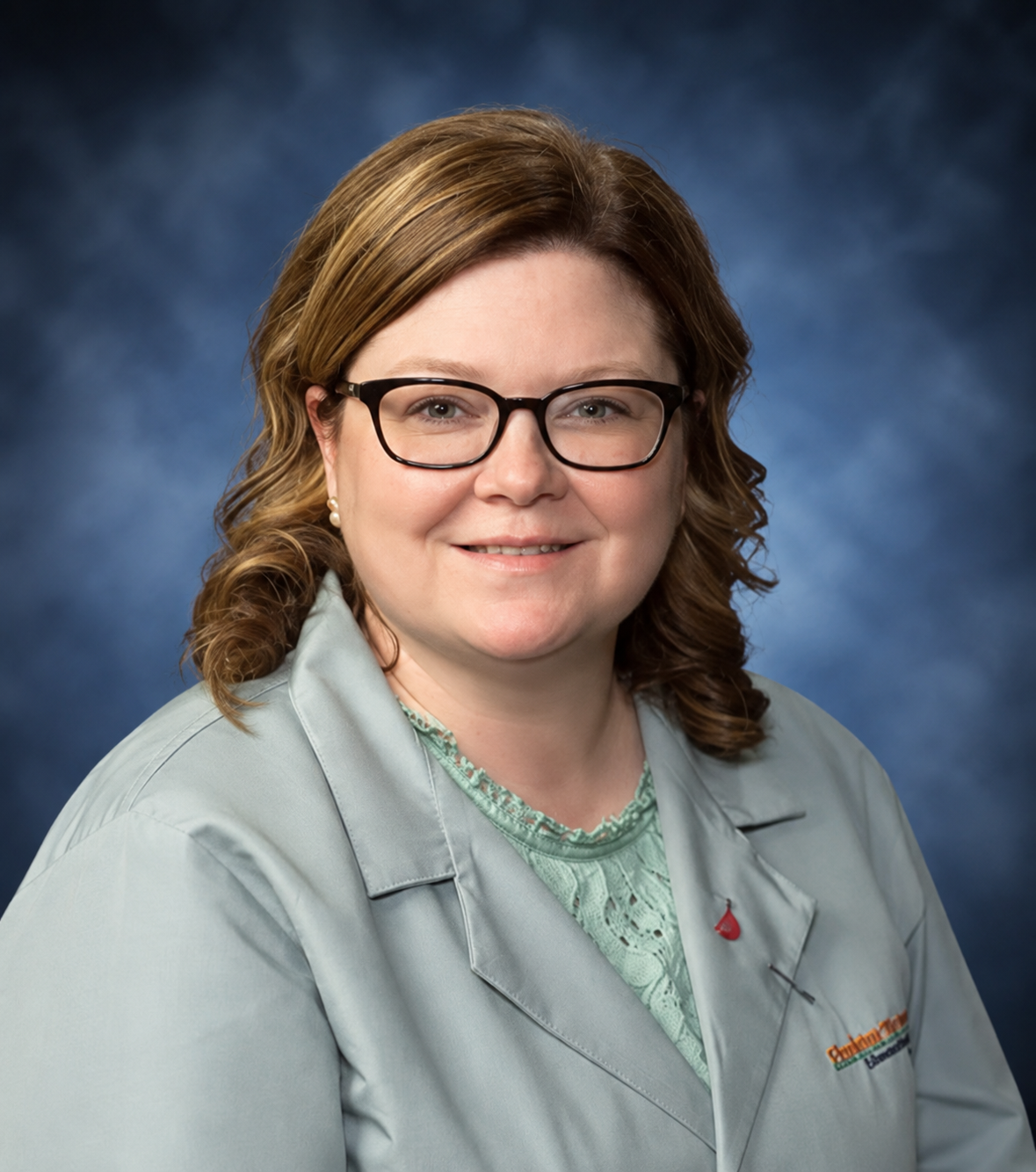 Courtney Harley, an APN with shoulder-length hair and glasses, wears a light-colored lab coat over a green top against a blue backdrop.